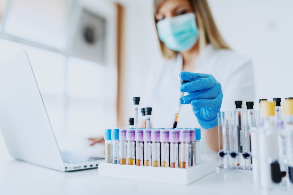 Close up of lab assistant in uniform, with mask and rubber gloves holding test tube with blood sample while sitting on chair and typing on laptop. Selective focus on test tubes.